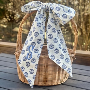 May include: A wicker basket with a blue and white floral patterned bow tied around the handle. The bow features an embroidered blue letter "S". The basket sits on a dark grey surface. The basket is made of woven wicker.