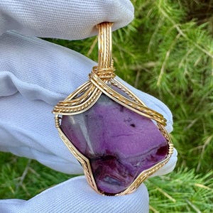 May include: A handcrafted pendant featuring a vibrant purple stone encased in a gold-colored wire wrap. The stone has a unique shape and texture, with a polished surface. The pendant is suspended from a matching gold-colored bail.