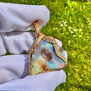 Könnte beinhalten: Ein golddrahtgewickelter Anhänger mit einem bunten Opal-Edelstein mit braunen Einschlüssen. Der Opal zeigt einen Regenbogen aus Farben, darunter Blau, Grün, Orange und Pink.