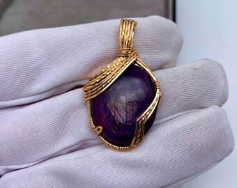 May include: A gold-colored pendant with a deep purple gemstone. The gemstone is faceted and encased in a wire-wrapped gold setting. The pendant is suspended from a gold bail.
