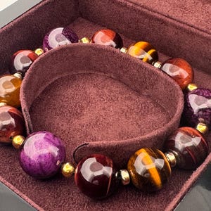 May include: A beaded bracelet with large, polished gemstone beads in shades of purple, brown, and gold. The bracelet is displayed inside a maroon jewelry box, showcasing the natural variations in the stones.