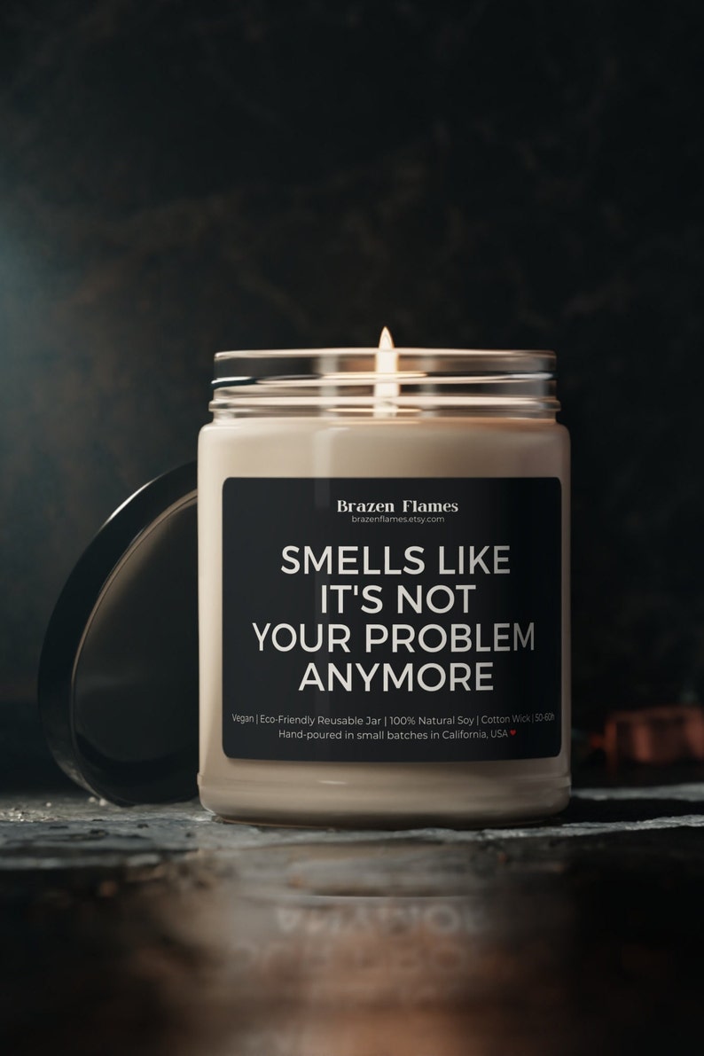 Puede incluir: Una vela encendida en un frasco transparente con una etiqueta negra. La etiqueta dice "SMELLS LIKE IT'S NOT YOUR PROBLEM ANYMORE". La vela est&aacute; hecha con cera de soja natural y una mecha de algod&oacute;n. Frasco reutilizable.