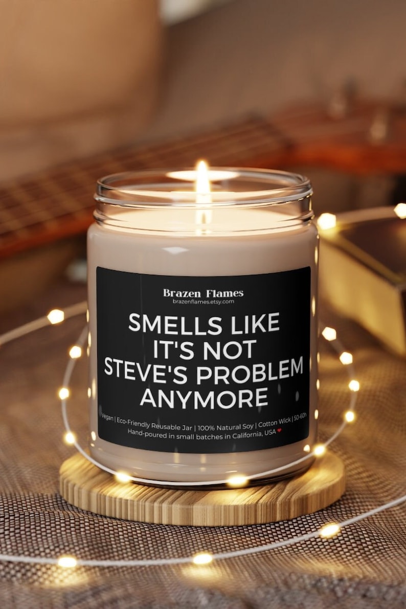 Puede incluir: Una vela encendida en un tarro de vidrio transparente con una etiqueta negra que dice "Smells Like It's Not Steve's Problem Anymore". La vela est&aacute; sobre un posavasos de madera, rodeada de luces de cadena.