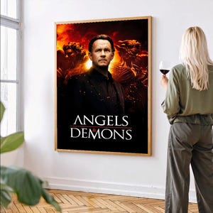 Könnte beinhalten: Ein gerahmtes Filmplakat für "Angels & Demons" mit Tom Hanks. Das Poster hat einen schwarzen Hintergrund mit weißem Text und einem feurigen Hintergrund. Das Poster befindet sich in einem Raum mit einem Fenster und einer Person, die ein Glas Rotwein hält.