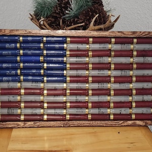 May include: A wooden framed American flag made from shotgun shells. The flag design uses blue, red, and silver shotgun shells to create the stripes and stars. The frame is a dark brown color.