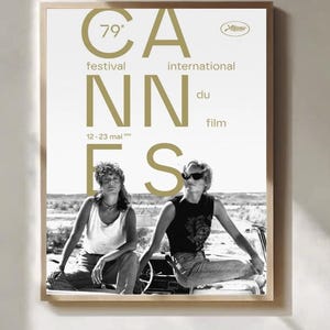 Op de afbeelding: Een ingelijste poster voor het 79e Internationale Filmfestival van Cannes. De poster toont een zwart-witfoto van twee mensen die op een auto zitten. De gouden tekst luidt "Cannes" en "12-23 mai".