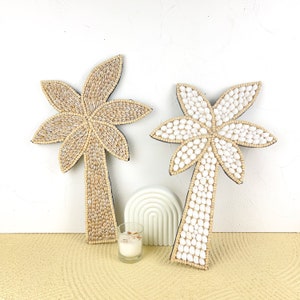 Könnte beinhalten: Zwei Palmen-Wanddekorationen aus natürlichen Materialien. Eine Palme ist weiß und die andere beige. Beide Palmen bestehen aus kleinen runden Perlen.