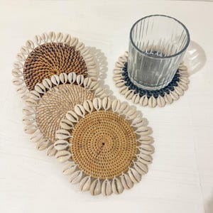May include: Four round woven coasters with a white shell trim. The coasters are made of natural materials and have a rustic look. A glass is sitting on one of the coasters.