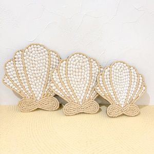 Op de afbeelding: Drie witte, schelpvormige wanddecoraties gemaakt van kleine witte kralen op een beige achtergrond.
