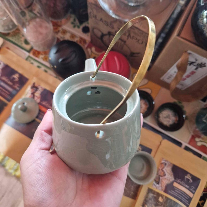 May include: A small, light green ceramic teapot with a gold-colored handle. The teapot has a round body and a small spout. The lid is off, revealing the interior. The background includes tea packaging and other tea-related items.