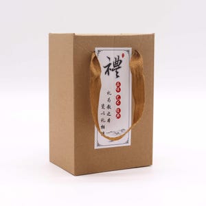 May include: A small, rectangular brown paper gift bag with a gold ribbon handle. A white label with red and black text is affixed to the front. The bag is designed for gifting.