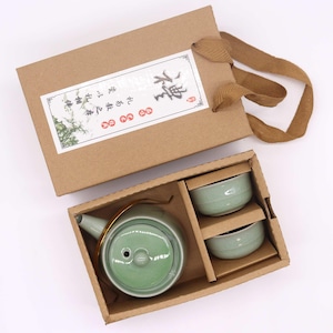 May include: A green ceramic tea set with a teapot and two cups, presented in a brown gift box with a handle. The teapot has a gold-colored handle. The box has a label with Asian characters.