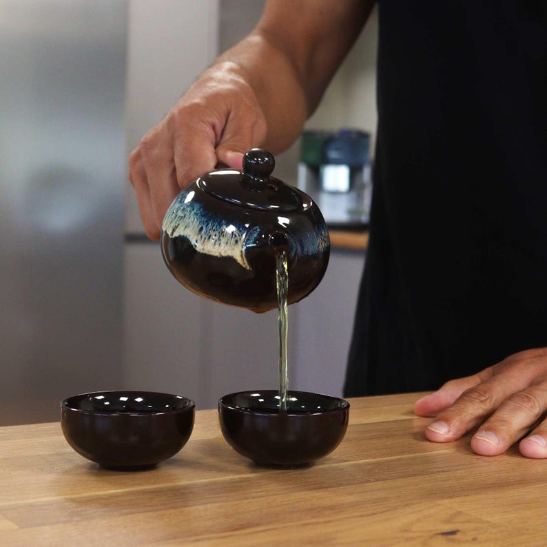 May include: A dark brown ceramic teapot with a blue and white glazed rim is pouring tea into two matching small bowls. The teapot has a round shape with a lid and a small handle. The tea is a light green color.