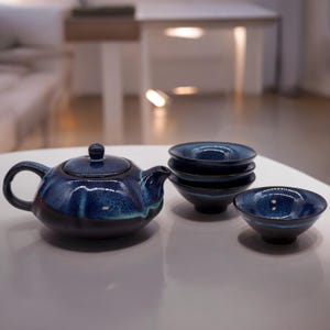Può includere: Servizio da tè in ceramica smaltata blu. Il set comprende una teiera con manico curvo e coperchio, e diverse tazze abbinate. Il servizio da tè è su una superficie bianca.