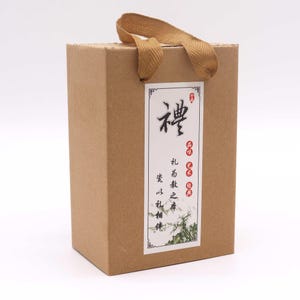 May include: A rectangular brown gift box with a tan handle. The box has a white label with Chinese characters and a decorative border. The handle is made of a woven tan material.