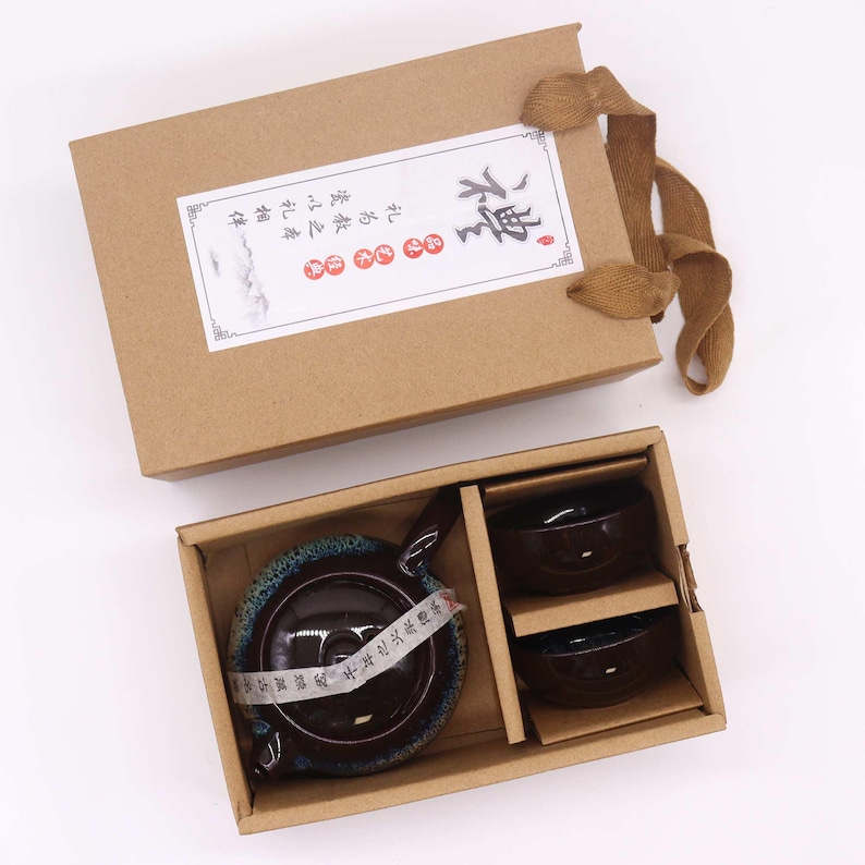 May include: A brown cardboard gift box with a tea set inside. The set includes a dark brown teapot with a blue rim and two matching cups. The box has a label with Chinese characters and a brown ribbon.
