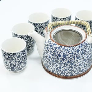 Könnte beinhalten: Ein weißes Keramik-Teeservice mit einer Teekanne und passenden Tassen. Die Teekanne hat einen geflochtenen Griff und einen Edelstahl-Infuser. Das Set hat ein blau-weißes Blumenmuster. Die Tassen sind zylindrisch.