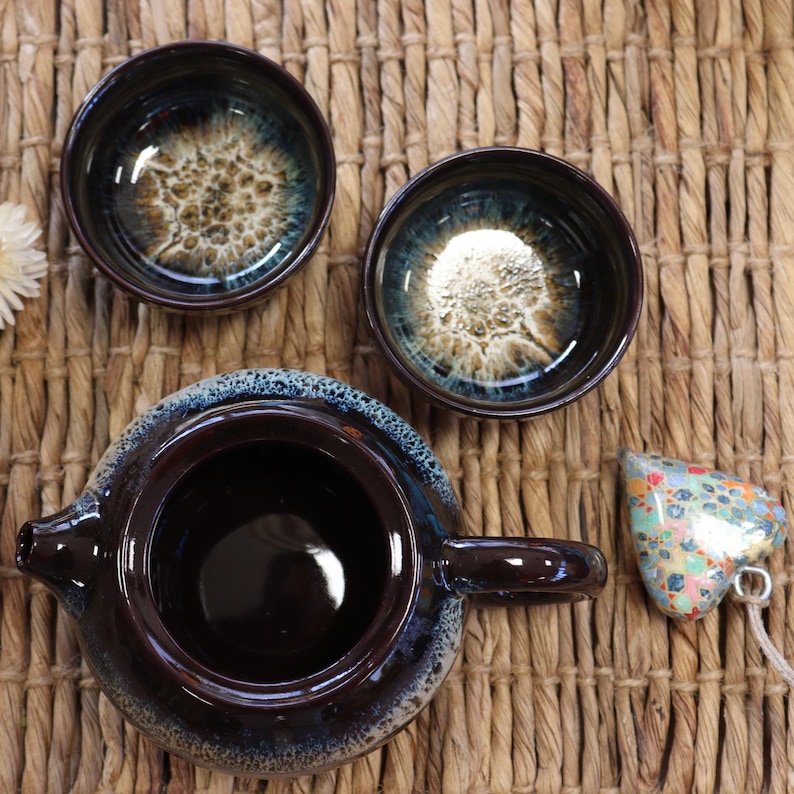 May include: A dark brown ceramic tea set with two small bowls and a teapot. The bowls and teapot have a blue and gold speckled interior. A heart-shaped charm with colorful patterns is also present.