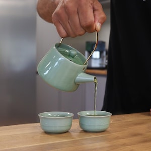 May include: A celadon green ceramic teapot with a gold handle is pouring tea into two matching cups. The tea set is on a wooden surface, suggesting a tea ceremony or a moment of relaxation.