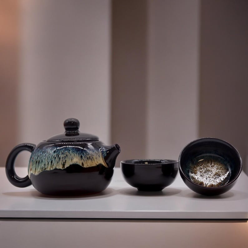 May include: A black ceramic teapot with a matching lid, handle, and spout, accompanied by two small black bowls. The teapot and bowls have a unique glaze with blue, tan, and gold tones. The set is designed for tea service.