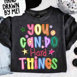May include: A dark gray t-shirt with the words "YOU CAN DO Hard THINGS" in colorful, bubble letters. The design includes stars, flowers, and a smiley face. The top left corner has a white speech bubble with the text "HAND DRAWN BY ME!"
