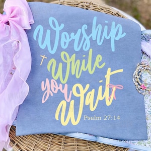 Peut inclure: Un t-shirt gris avec les mots "Worship While You Wait" en écriture bleue, verte, rose et jaune. Le texte "Psalm 27:14" est en dessous. Un nœud rose et un bracelet sont visibles.