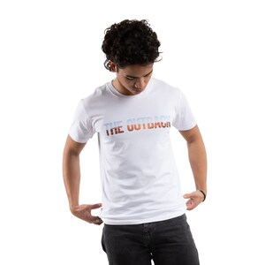 May include: White t-shirt featuring the text "THE OUTBACK" in a gradient of blue, orange, and brown. The short-sleeved shirt is likely cotton. The model is wearing dark grey jeans.