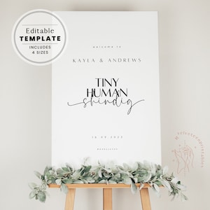 May include: A white printable sign with black text that reads "Welcome to Kayla & Andrews Tiny Human Shindig 16.09.2023 #babyjones". The sign is on a wooden easel with green foliage in front of it.