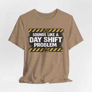 May include: A brown t-shirt with the text "SOUNDS LIKE A DAY SHIFT PROBLEM" in white, with yellow and black hazard stripes and warning symbols. The shirt is made of a soft material.