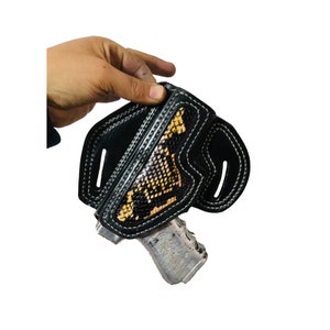 Puede incluir: Funda de cuero negro con un adorno de estampado de piel de serpiente. La funda está cosida con hilo blanco y diseñada para sujetar una pistola. El estampado de piel de serpiente presenta tonos de marrón, beige y amarillo.