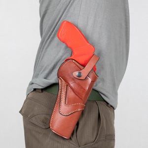 May include: A brown leather holster with a bright orange handgun. The holster is worn on the hip and features decorative stitching and a strap. The handgun's grip is visible above the holster.
