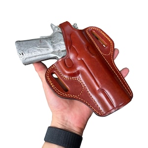 Puede incluir: Una funda de cuero marrón con una pistola plateada. La funda está cosida con un hilo marrón más claro y tiene un acabado brillante. El arma de fuego está parcialmente insertada en la funda, mostrando su forma y diseño. La imagen está sobre un fondo blanco.