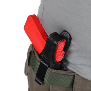 Puede incluir: Una funda de cuero negra con una pistola de plástico roja en su interior. La funda está unida a un cinturón verde.