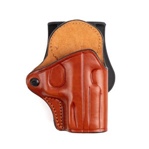 May include: A brown leather holster with a black clip. The holster has a smooth, polished finish and visible stitching. The interior is a lighter tan colour. The holster is designed to hold a handgun.