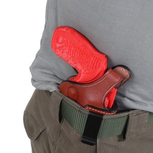 Könnte beinhalten: Eine rote Pistole in einem braunen Lederholster mit einem schwarzen Clip, der an einem grünen Gürtel befestigt ist.