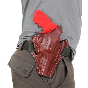 Op de afbeelding: Een bruine leren holster met een rood plastic pistool erin. De holster is bevestigd aan een groene riem.