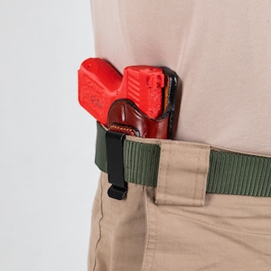 Könnte beinhalten: Eine rote Pistole in einem braunen Lederholster, das an einem schwarzen Gürtelclip befestigt ist. Das Holster ist an einem grünen Gürtel befestigt.