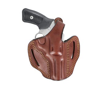 Op de afbeelding: Een bruin leren holster met een revolver. De holster heeft decoratieve stiksels en uitsparingen. De revolver heeft een zwarte grip en een zilveren loop.
