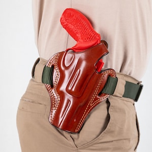 Könnte beinhalten: Ein braunes Lederholster mit einer roten Pistole darin. Das Holster ist an einem grünen Gürtel befestigt.