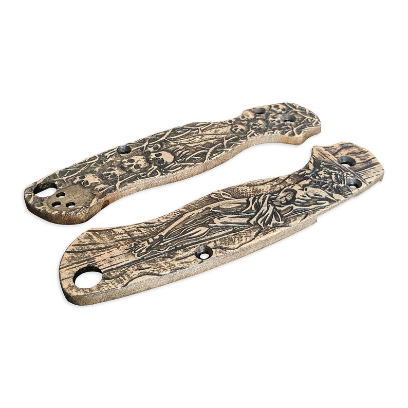 May include: Two brown knife scales with intricate engraved designs. One scale features a skull and crossbones motif, while the other depicts a figure on a cross.