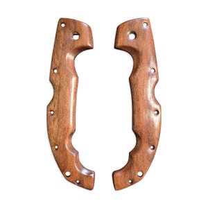 Può includere: Due manici di coltello in legno non finiti con una macchia marrone. I manici hanno una forma curva e diversi fori praticati.
