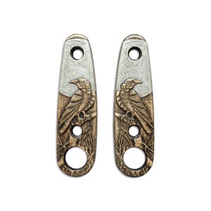 Könnte beinhalten: Zwei dekorative, längliche Metallstücke mit einem Rabenmuster. Jedes Stück hat einen silbernen oberen Abschnitt, einen geschnitzten Raben auf einem Ast und einen unteren Abschnitt mit einem kreisförmigen Loch. Die Raben sind mit Braun- und Beigetönen detailliert.