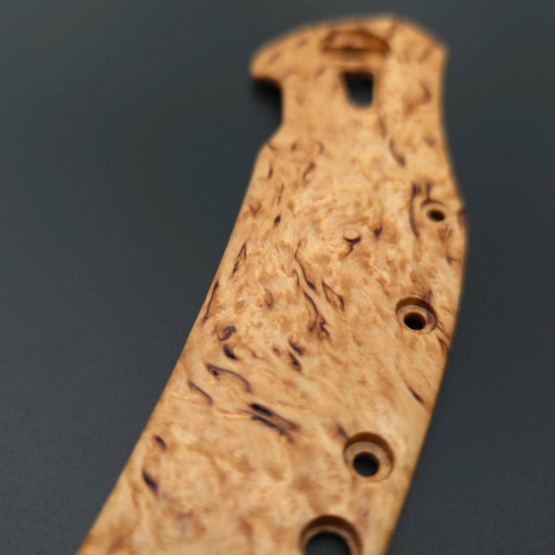 Puede incluir: Primer plano de un mango de cuchillo de madera clara con vetas marrones oscuras. El mango tiene una forma curva con tres agujeros circulares y un recorte cerca de la parte superior. El patr&oacute;n de la veta de la madera es visible.