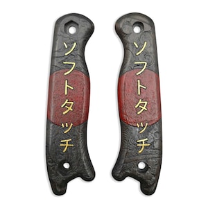 May include: Two dark gray knife handles with a red center band and gold Japanese text. The handles have a textured pattern and a hole at the top for attachment. The Japanese text reads "Soft Touch".