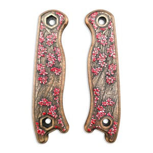 May include: Pair of decorative bronze-colored knife handle scales with a floral cherry blossom design in red and white. The scales have a textured wood-grain appearance and hexagonal and circular holes for mounting.