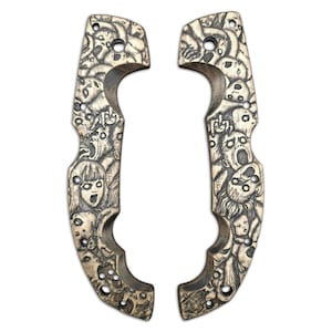Può includere: Due guancette per coltello in legno con un disegno intagliato di volti urlanti. Le guancette sono marrone chiaro con un disegno intagliato marrone scuro. Hanno fori per viti e una forma curva dell'impugnatura.