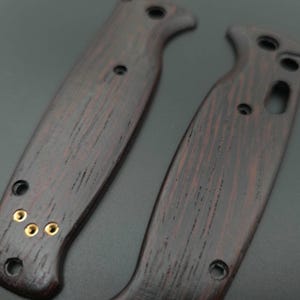 Pu&ograve; includere: Due guancette per manico di coltello in legno marrone scuro con venature visibili e rivetti in ottone. Le guancette hanno una forma curva con un'incavatura per le dita e diversi fori preforati. Il legno ha una tonalit&agrave; rossastra.