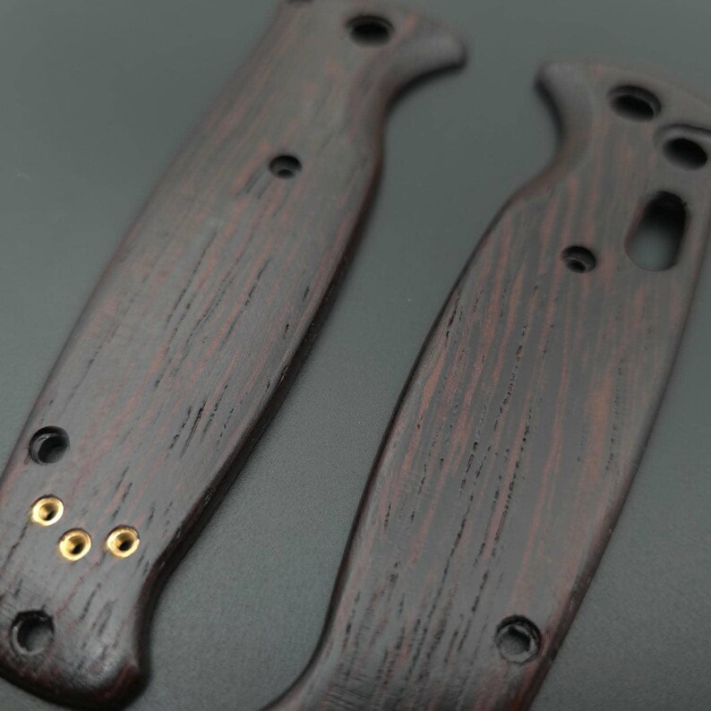 Pu&ograve; includere: Due guancette per manico di coltello in legno marrone scuro con venature visibili e rivetti in ottone. Le guancette hanno una forma curva con un'incavatura per le dita e diversi fori preforati. Il legno ha una tonalit&agrave; rossastra.