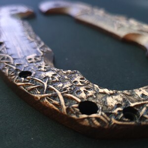 May include: Close-up of a bronze-colored horseshoe with intricate carvings. The horseshoe features a detailed design with skulls and floral patterns. The metal has a textured surface and three visible holes.