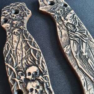 May include: Two intricately carved wooden knife handles. One handle features a skull and crossbones design with a dark brown finish. The other handle features a detailed carving of a figure with a crown of thorns, with a lighter brown finish.